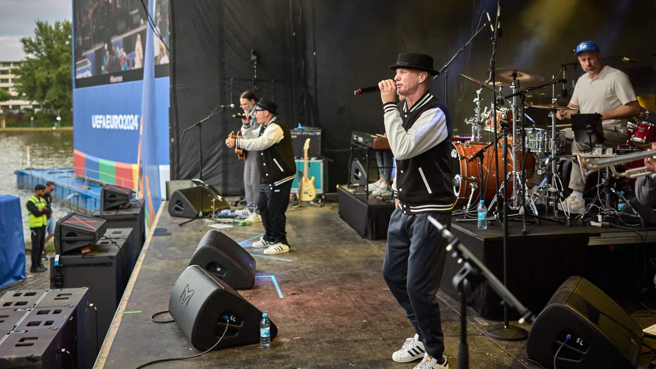 Musiker stehen lässig mit Hut und Collegejacke auf einer Bühnen und singen