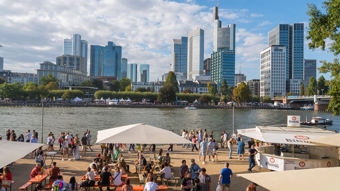 Menschen sitzen an Bierbänken unter Sonnenschirmen am Mainufer, mit Frankfurter Skyline und Besucher*innen im Hintergrund.