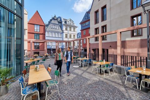 A couple walks on a cobbled square in Frankfurt's old town, surrounded by colourful, historic buildings. In the foreground are tables and chairs in an outdoor area of a restaurant.