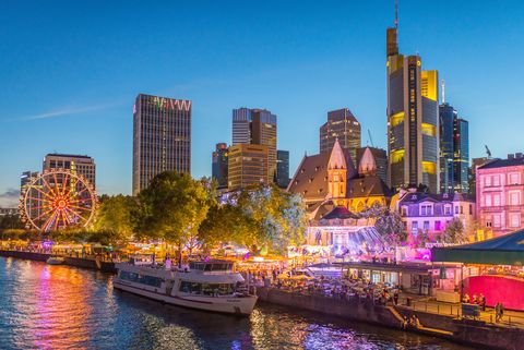 Festively illuminated Main Festival with Ferris wheel, carousel, Frankfurt skyline and crowds of people on the riverbank in the evening.