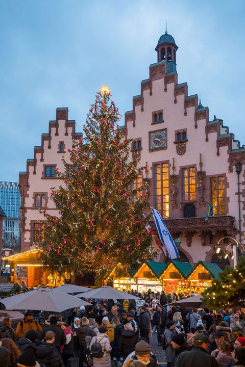 Beleuchteter Weihnachtsbaum auf dem Frankfurter Weihnachtsmarkt 2023 vor der historischen Kulisse des Römers, mit geschmückten Marktständen und vielen Menschen. Das Bild zeigt eine festliche Atmosphäre in der Abenddämmerung. 