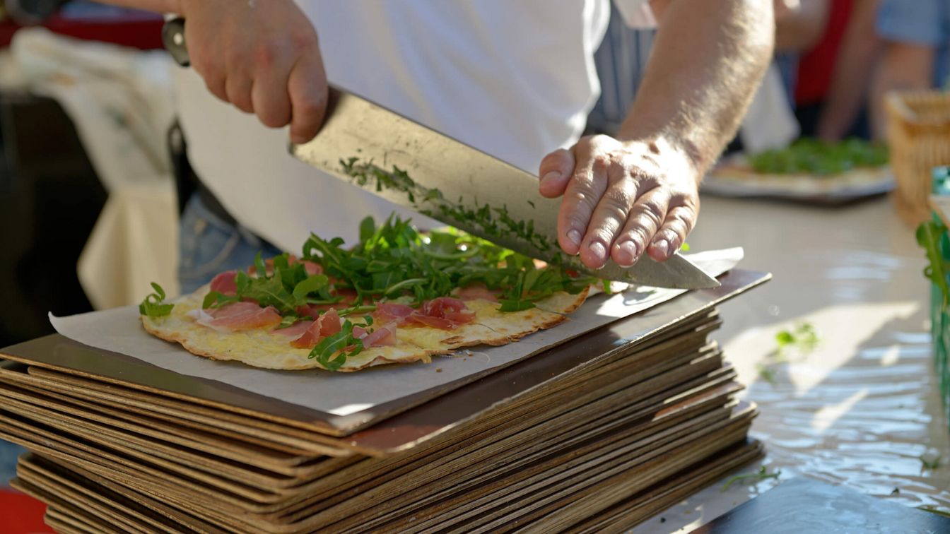 Nahaufnahme eines Flammkuchens, der mit Rucola und Schinken belegt ist. Eine Person schneidet den Flammkuchen mit einem großen Messer, während er auf gestapelten Blechen liegt.