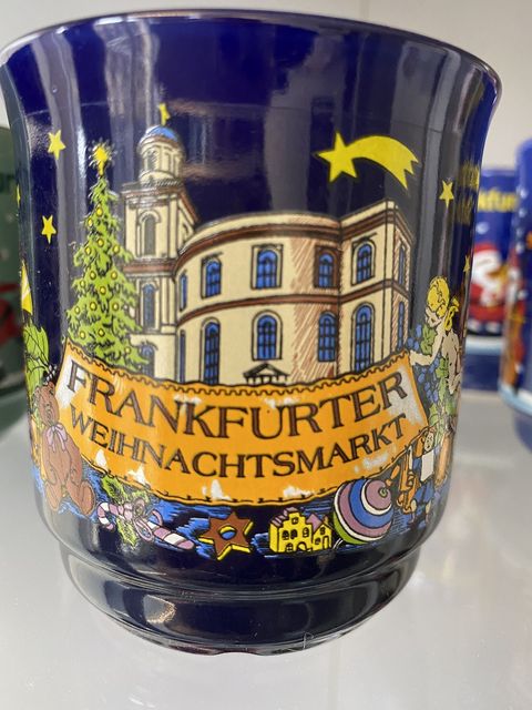 Dunkelblaue Glühweintasse mit einer Darstellung der Paulskirche. Im Vordergrund ein Banner, gehalten von einem geschmückten Weihnachtsbaum und einem Engel, mit der Aufschrift Frankfurter Weihnachtsmarkt. Vor dem Banner ist ein Zimtstern, eine Zuckerstange, ein Teddybär sowie ein Kinderkreisel abgebildet. Gekrönt ist die Abbildung von einem Weihnachtsstern. 