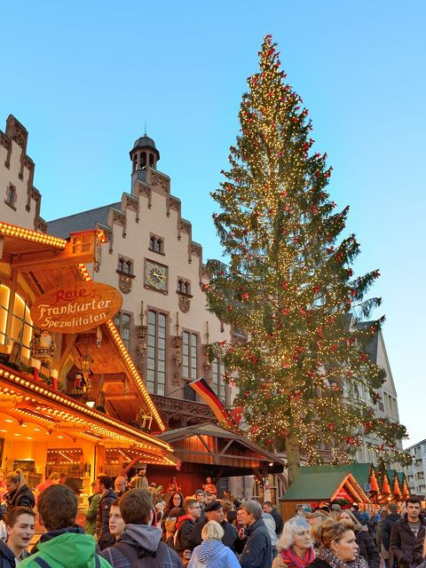 Beleuchteter Weihnachtsbaum auf dem Frankfurter Weihnachtsmarkt 2015 vor historischer Kulisse des Römers, mit geschmückten Marktständen und Personen im Vordergrund. Das Bild zeigt eine festliche Atmosphäre am Abend. 