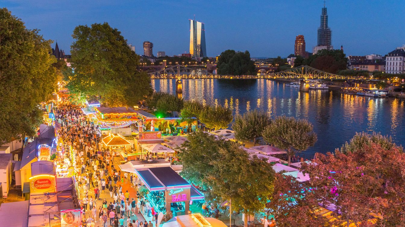 Marktstände und bunt beleuchtete Buden am Mainufer, mit Menschenmassen und Brückenlichtern bei Abendstimmung.