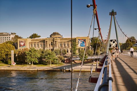 View of the Städel Museum in Frankfurt with its magnificent exterior façade, surrounded by trees, in the foreground the Holbeinsteg with walkers over the Main.