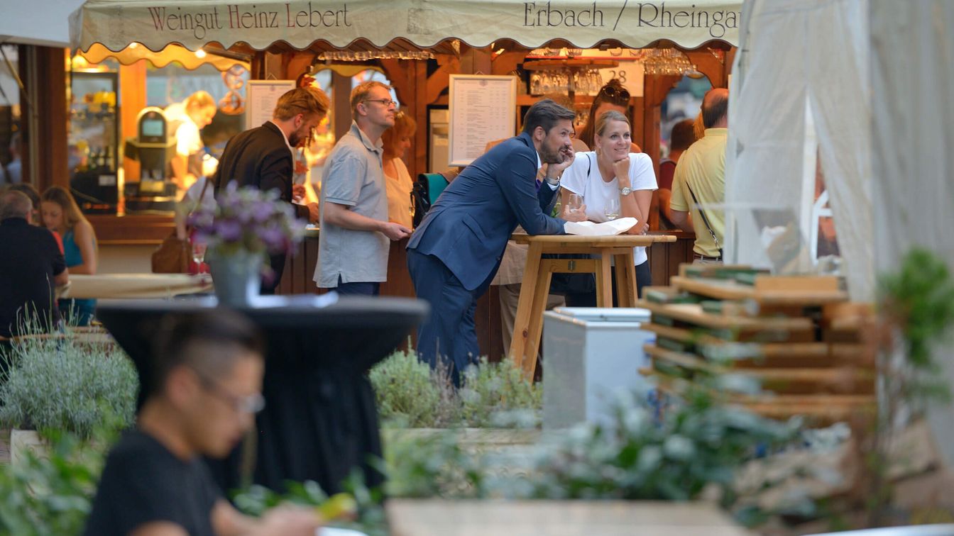 A group of people stand and chat at a wine bar with rustic charm. The Rheingau wine market has a convivial atmosphere, surrounded by festively decorated stalls and a summery ambience.