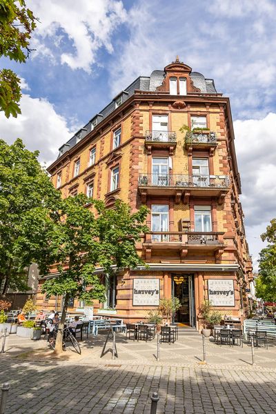 Historic building with balconies and café on the ground floor, surrounded by trees and street flair.