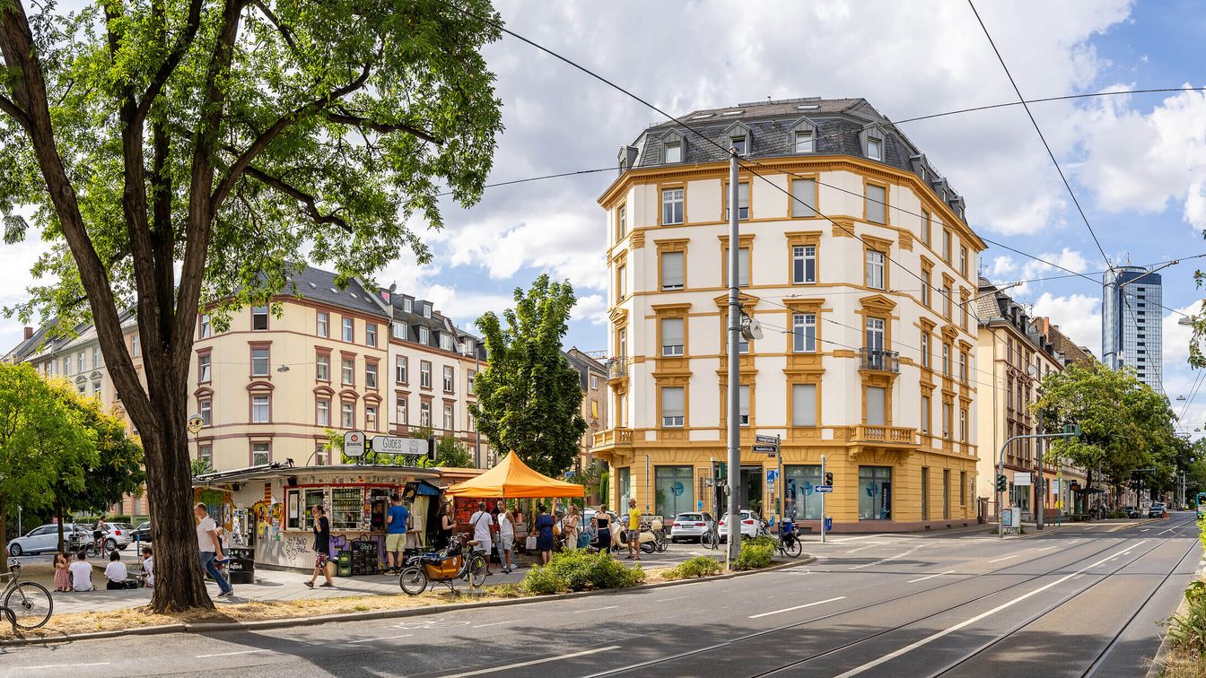 Eine Kreuzung an der Friedberger Landstraße im Frankfurter Nordend, flankiert von historischen Gebäuden und Bäumen. Menschen genießen den Sommer in einem kleinen Verkaufsstandbereich.