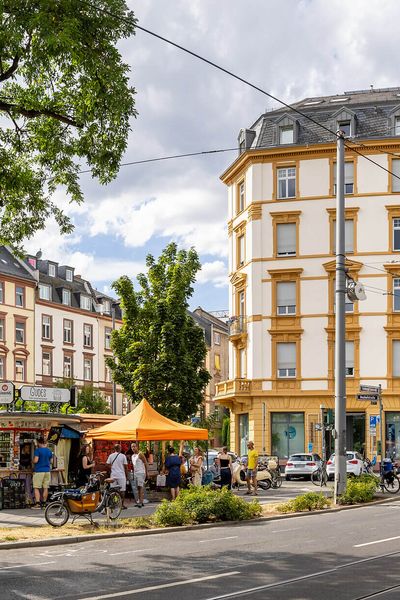 A crossroads on Friedberger Landstraße in Frankfurt's Nordend district, flanked by historic buildings and trees. People enjoy the summer in a small stall area.