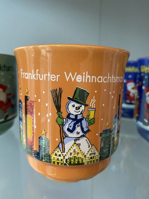 Die Glühweintasse ist in orange. Auf der Tasse ist die Frankfurter Skyline sowie die historischen Gebäude der Stadt zu sehen. In der Mitte ist ein traditioneller Schneemann mit heißem Apfelwein sowie mit einem Besen dargestellt. Oben am Rand der Tasse steht Frankfurter Weihnachtsmarkt. 