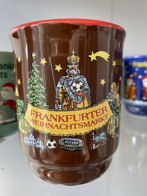 Eine braune Glühweintasse mit der Darstellung eines Kaisers, der anstelle des Reichsapfel ein Fußball hält. Darunter ein Banner gehalten von einem Engel und dem Weihnachtsbaum mit der Aufschrift Frankfurter Weihnachtsmarkt. Unter dem Banner abgebildet ist das Frankfurter Fußball Stadion, der Frankfurter Dom, ein Fußball und der Frankfurter Römer. Oben rechts befindet sich der Weihnachtsstern. . . 