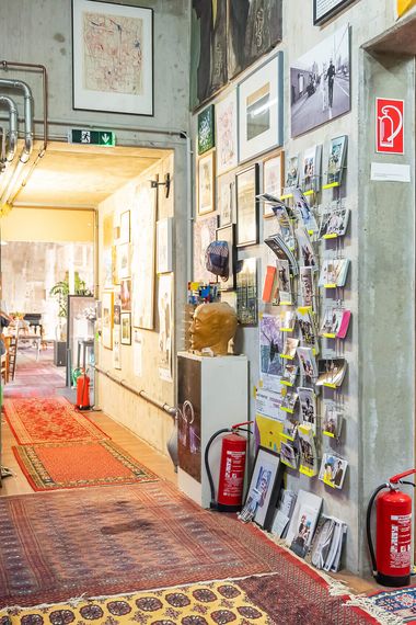 Kreatives Atelier mit bunten Teppichen, gerahmten Bildern an Wänden, Kunstwerken, orangem Sofa, Fahrrad und Feuerschutzmitteln, voller künstlerischer und gemütlicher Details.