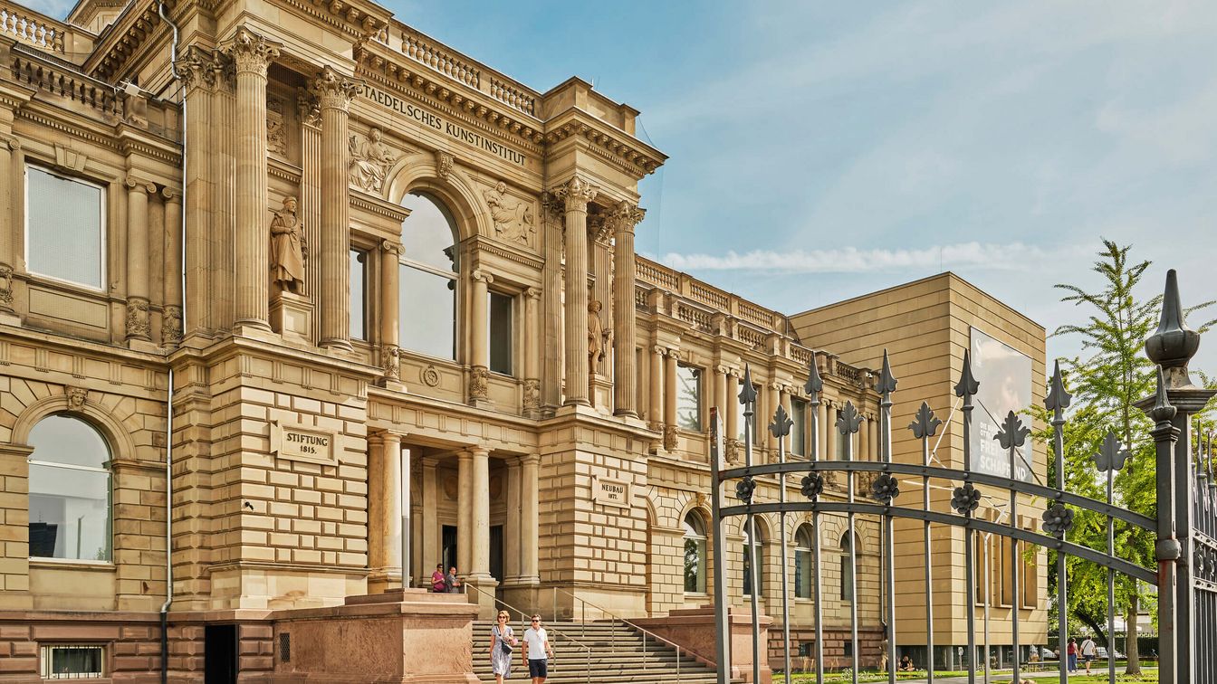 Das Städel Museum in Frankfurt am Main an einem sonnigen Tag. Das historische, neoklassizistische Gebäude mit Säulen und kunstvoller Fassade wird von einem schmiedeeisernen Zaun im Vordergrund eingerahmt. Einige Besucher*innen sind vor dem Eingang zu sehen. Rechts steht ein moderner Erweiterungsbau