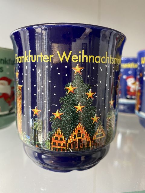 Dunkelblaue Glühweintasse mit einem festlich geschmückten Weihnachtsbaum. Auf dem Baum sind Sternen prominent platziert. Vor dem Weihnachtsbaum ist der Frankfurter Römer sowie die Skyline zu erkennen. Es schneit in der Abbildung und über dem Weihnachtsbaum steht in gelber Schrift Frankfurter Weihnachtsmarkt. 